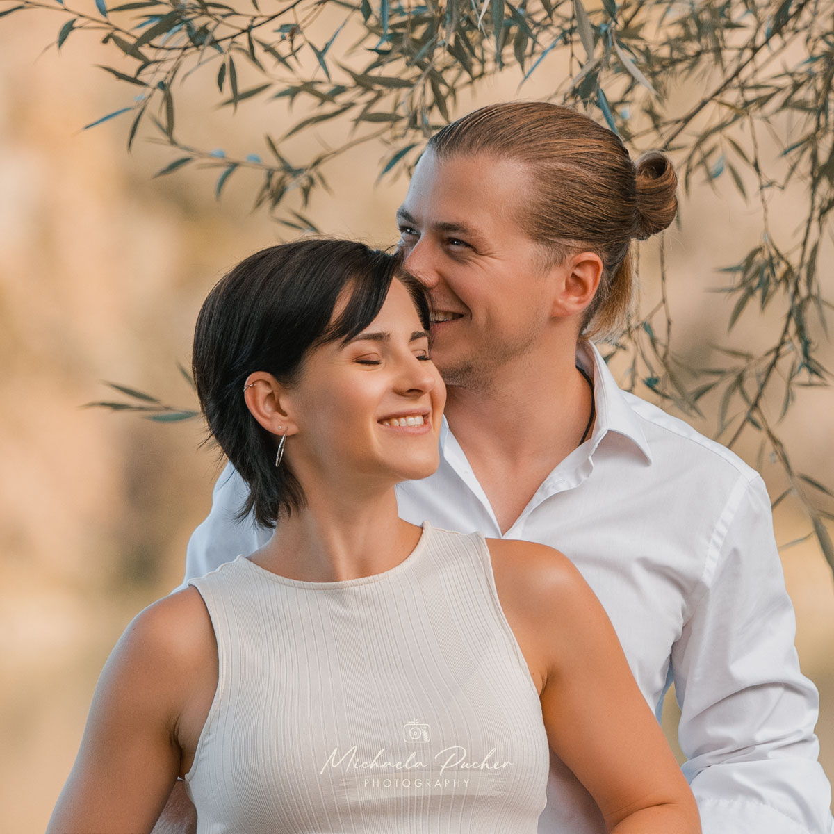 Ein Paar steht eng umeinander geschlungen; die Frau lächelt entspannt mit geschlossenen Augen, während ihr Partner liebevoll hinter ihr lächelt.